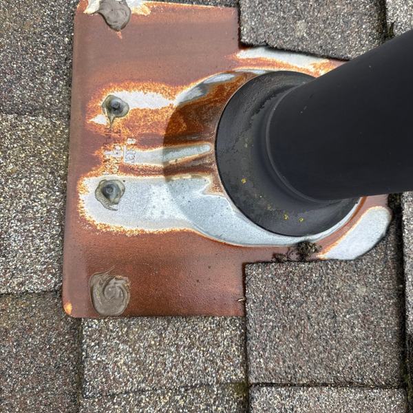 A vent pipe passing through a roof, with the metal flashing plate around it heavily rusted and the sealant cracked