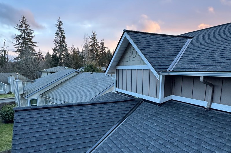 A finished dark grey asphalt shingle roof on a two storey house at dusk, with a gabled dormer and evergreens behind it