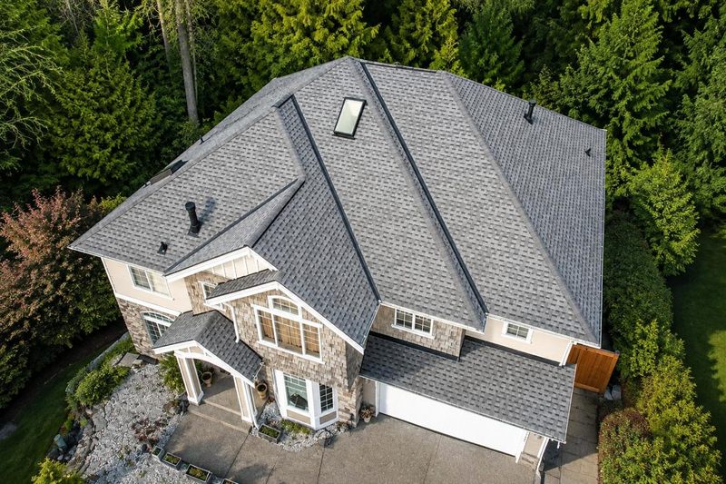 Aerial view of a completed grey architectural shingle roof on a two storey house, clean hip and ridge lines, a skylight and a new copper coloured gutter, evergreens on every side