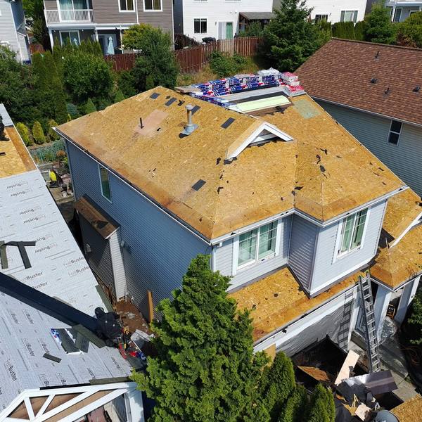 Aerial view of a house re-roof in progress, the main roof decked in fresh sheathing, underlayment already down on the section alongside and new shingle bundles stacked ready